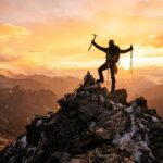 A lone climber standing on a rocky mountain summit at sunrise.